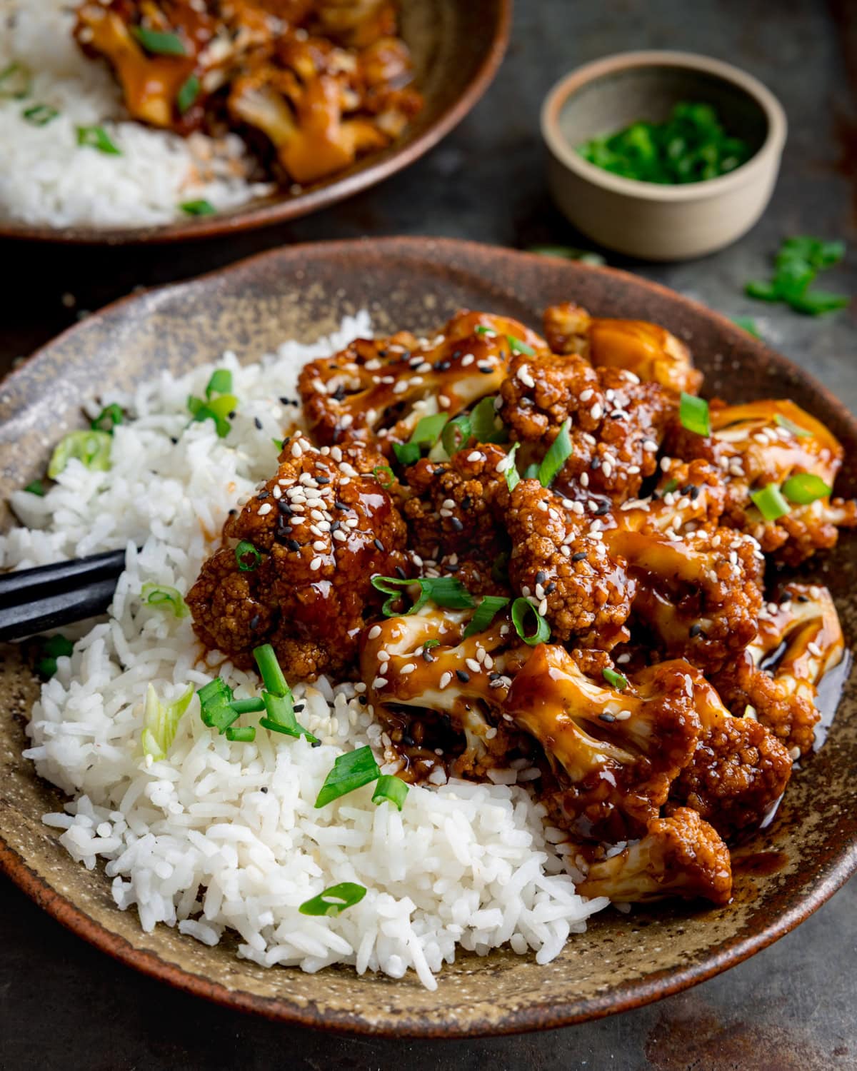 A bowl of white rice topped with sesame cauliflower in a dark sticky sauce, garnished with chopped spring onions, with a small dish of spring onions in the background.