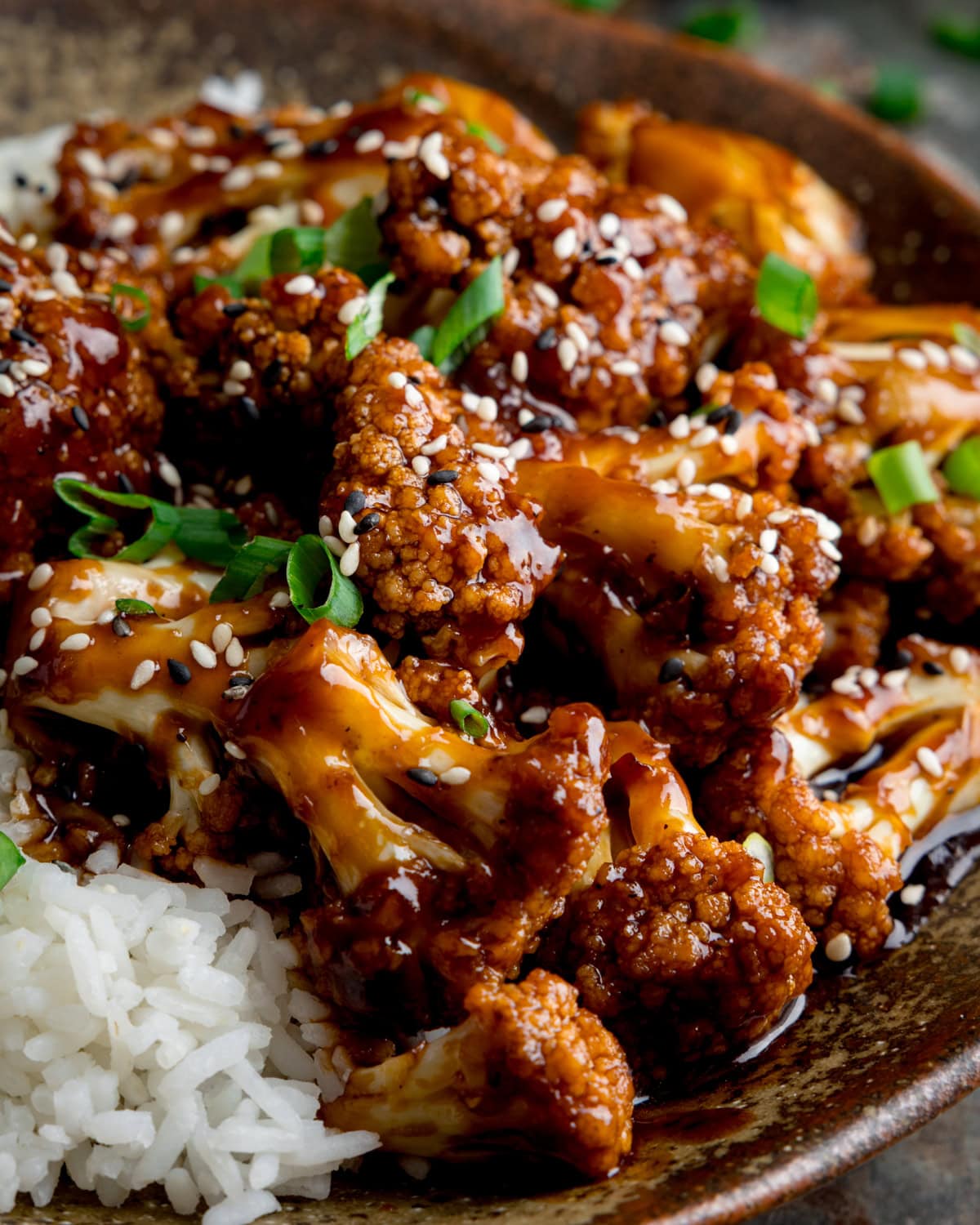 Close-up of glazed sesame cauliflower florets topped with sesame seeds and sliced spring onions, served with white rice in a brown bowl.