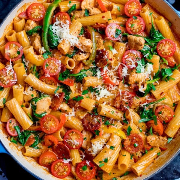 A frying pan filled with rigatoni pasta, cherry tomatoes, spinach, and tender Cajun chicken pieces, topped with herbs and grated cheese.