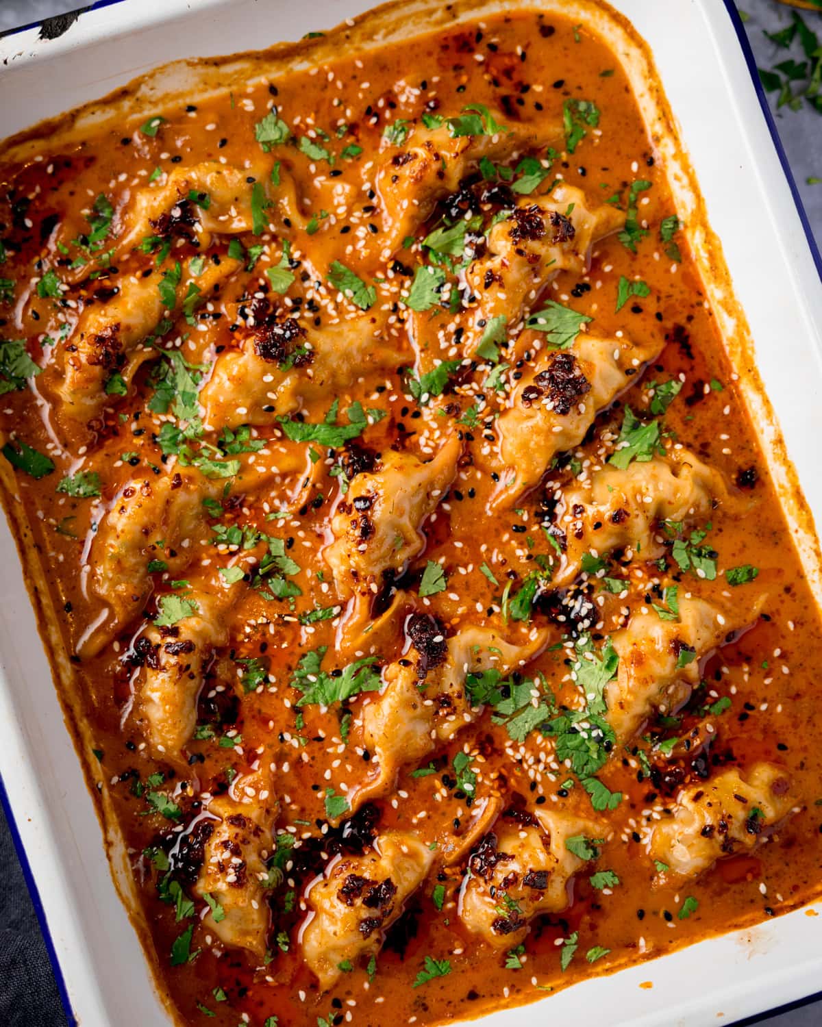 A tray of gyoza dumplings covered in a rich, orange-red curry sauce, garnished with chopped coriander, sesame seeds, and chilli oil.