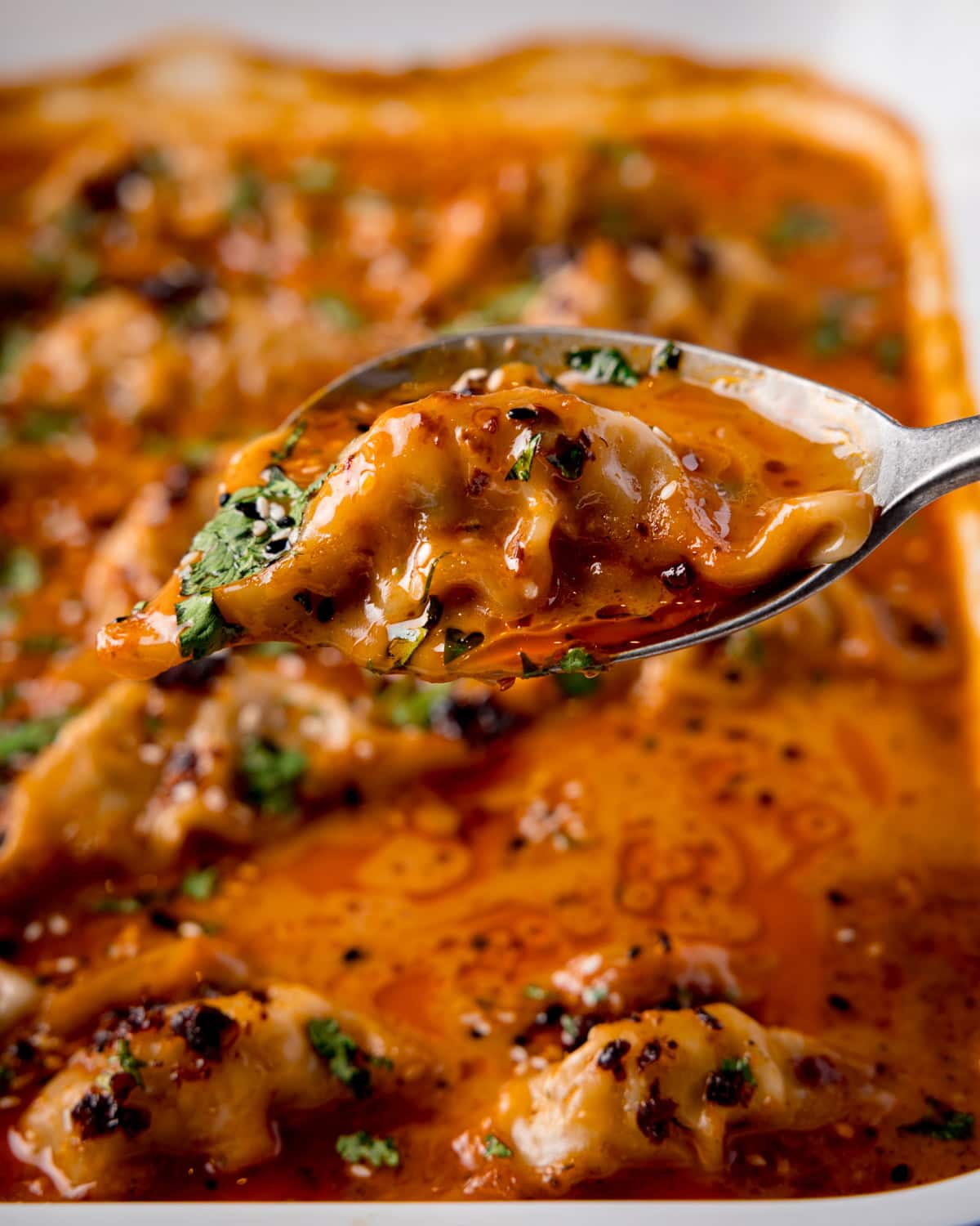 A close-up of a spoon lifting dumplings covered in thai red curry sauce, garnished with chopped herbs, from a tray.