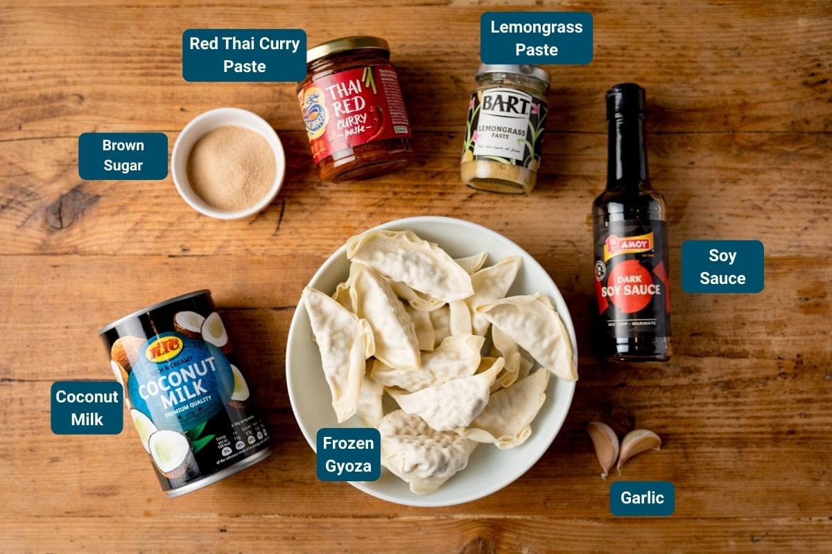 A bowl of frozen gyoza surrounded by labelled ingredients: coconut milk, brown sugar, red Thai curry paste, lemongrass paste, soy sauce, and garlic on a wooden surface.