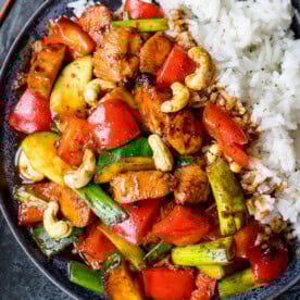 A plate of kung pao chicken with red peppers, courgette, spring onions, cashews, and white rice.