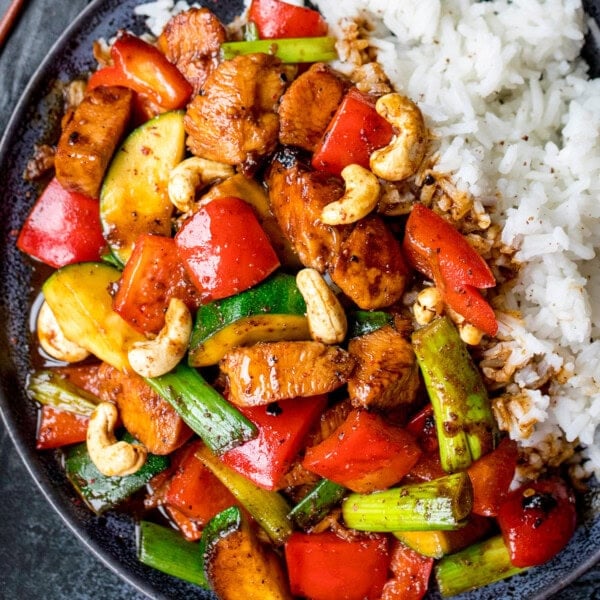 A plate of kung pao chicken with red peppers, courgette, spring onions, cashews, and white rice.