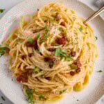 An overhead image of a plate of spaghetti carbonara topped with grated cheese, crispy bacon pieces, chopped parsley, and black pepper, served on a white plate with a fork.