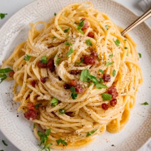 An overhead image of a plate of spaghetti carbonara topped with grated cheese, crispy bacon pieces, chopped parsley, and black pepper, served on a white plate with a fork.