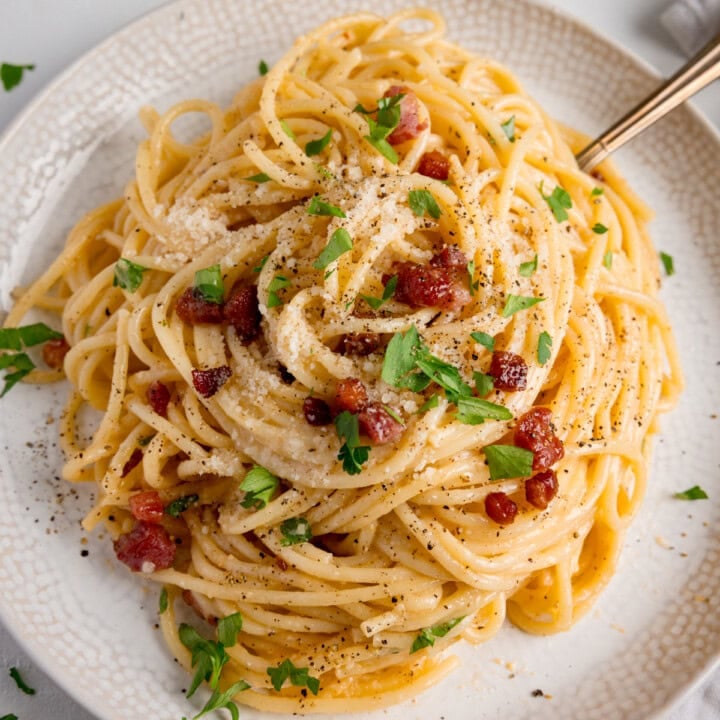 An overhead image of a plate of spaghetti carbonara topped with grated cheese, crispy bacon pieces, chopped parsley, and black pepper, served on a white plate with a fork.