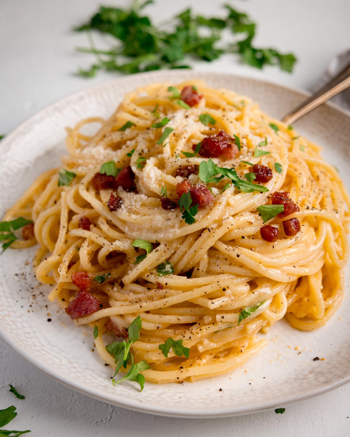 A white plate of spaghetti carbonara topped with pancetta, grated cheese, black pepper, and fresh parsley, with a fork at the side. The plate is on a wide background.
