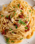 Close up overhead of a plate of spaghetti carbonara topped with grated cheese, crispy pancetta pieces, chopped parsley, and black pepper.
