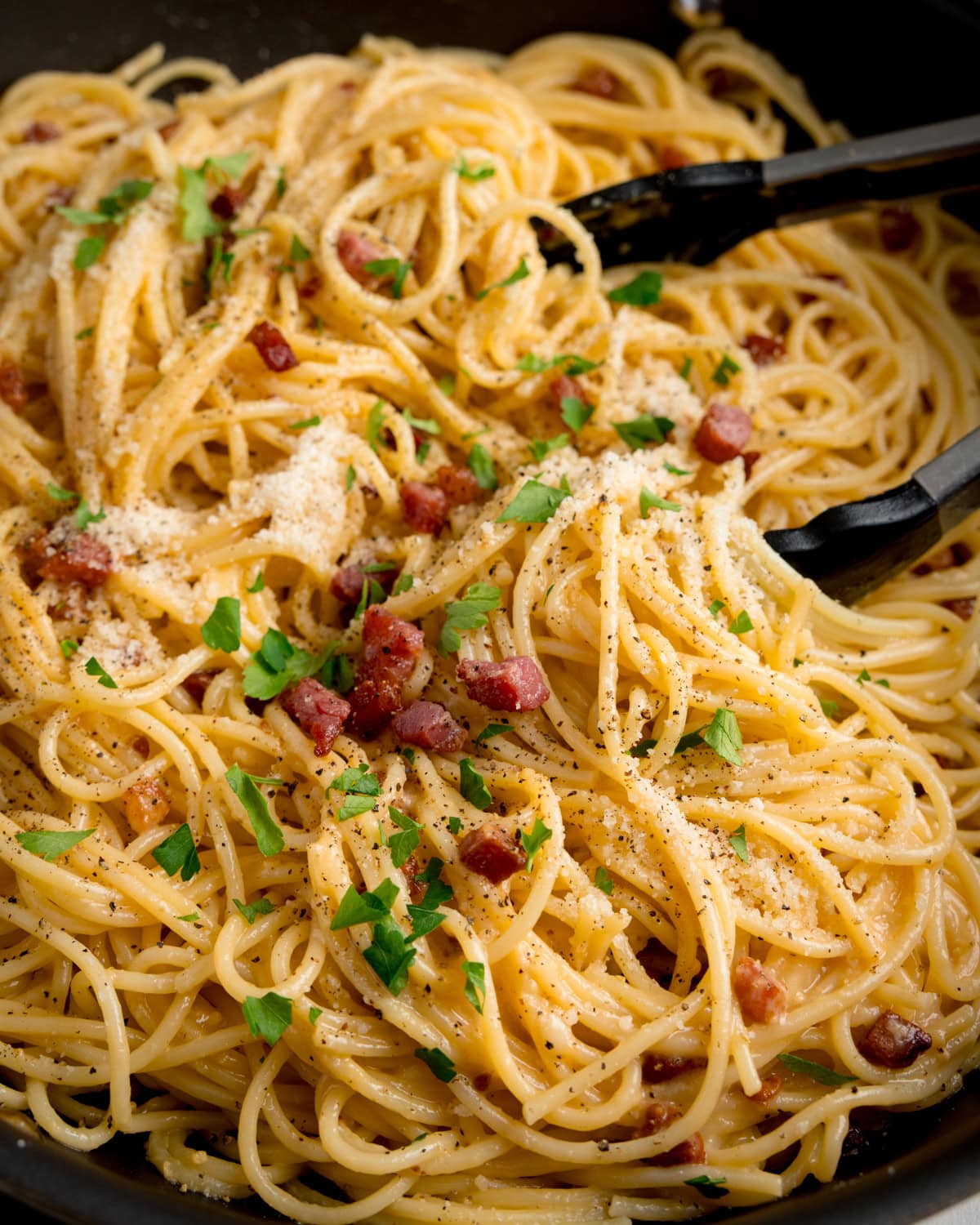 A close-up of spaghetti carbonara in a pan, topped with chopped parsley, grated cheese, pancetta, and black pepper, with black serving tongs on the side.