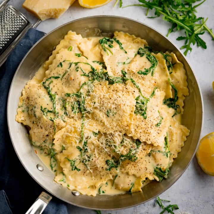A frying pan filled with ravioli in a creamy lemon sauce, topped with grated cheese and rocket, with lemon wedges and herbs on the side.