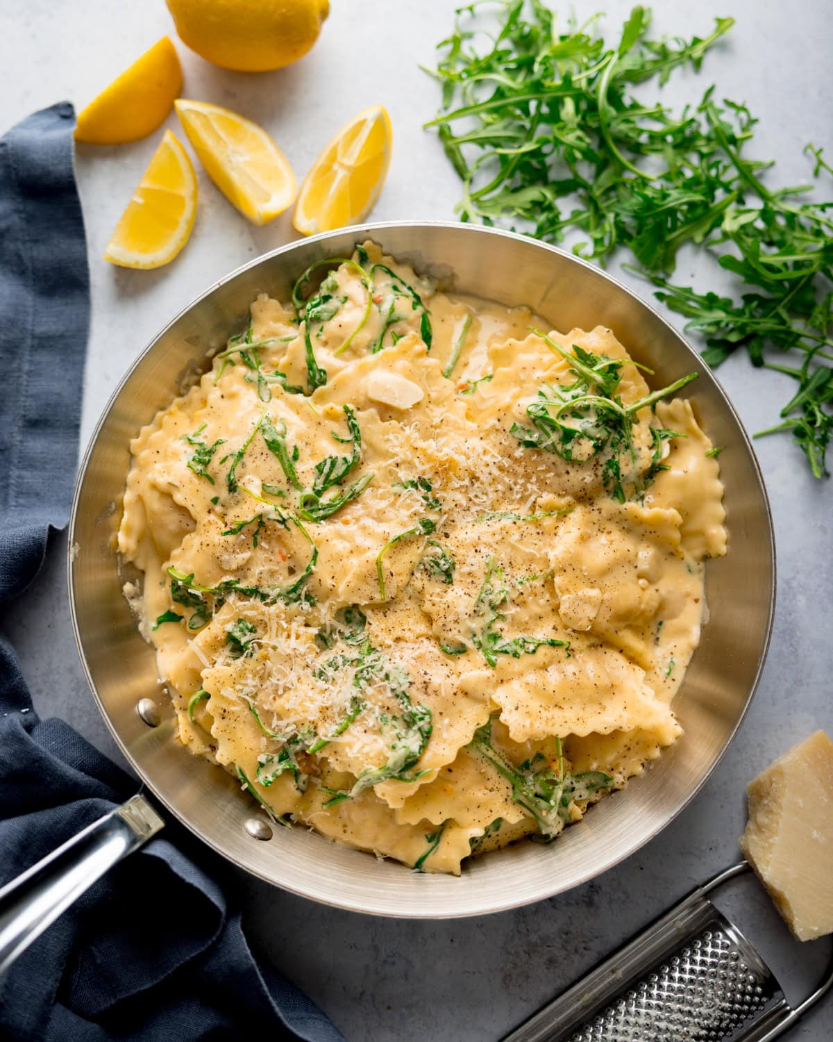 A stainless steel pan filled with creamy lemon ravioli topped with grated cheese and rocket, surrounded by lemon wedges, rocket leaves, and a cheese grater.