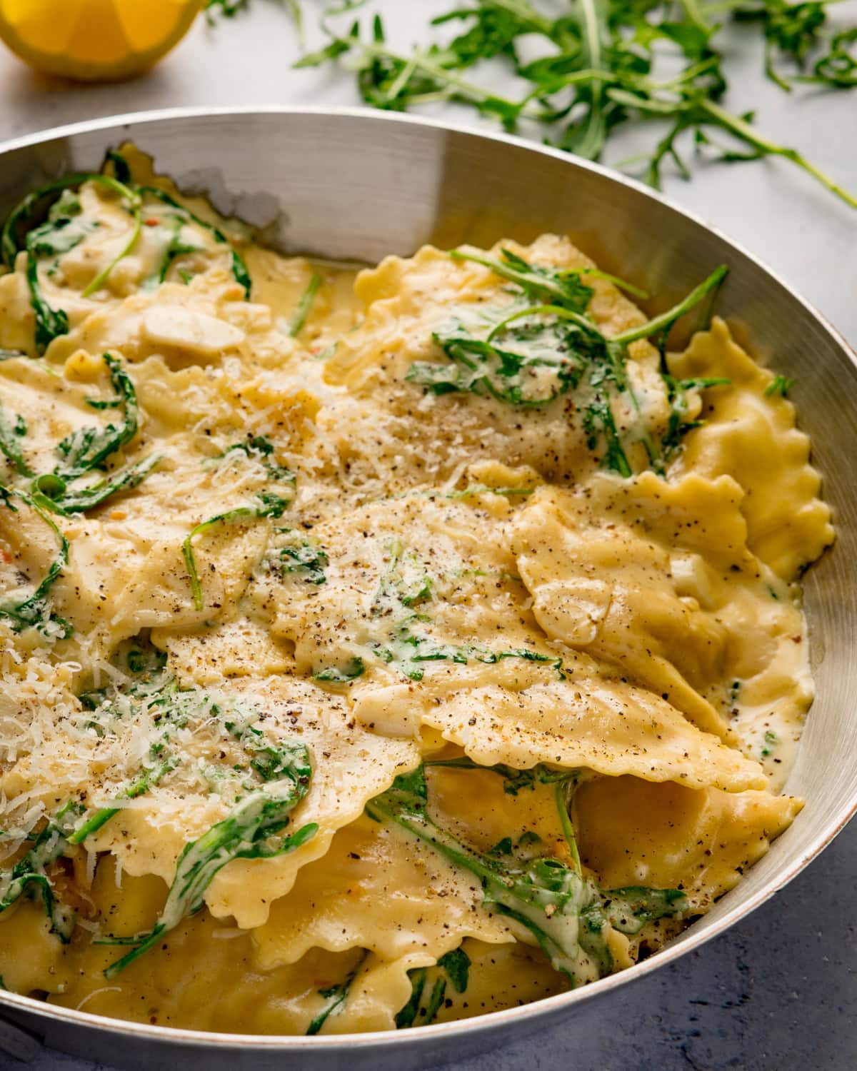 A stainless frying pan full of creamy lemon ravioli, topped with grated cheese, black pepper, and fresh rocket.