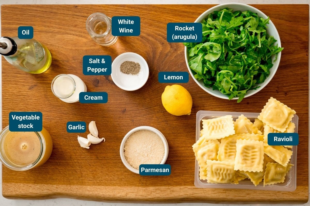 A wooden board displays ingredients for a pasta dish: oil, vegetable stock, garlic, cream, salt and pepper, white wine, parmesan, lemon, rocket and ravioli.