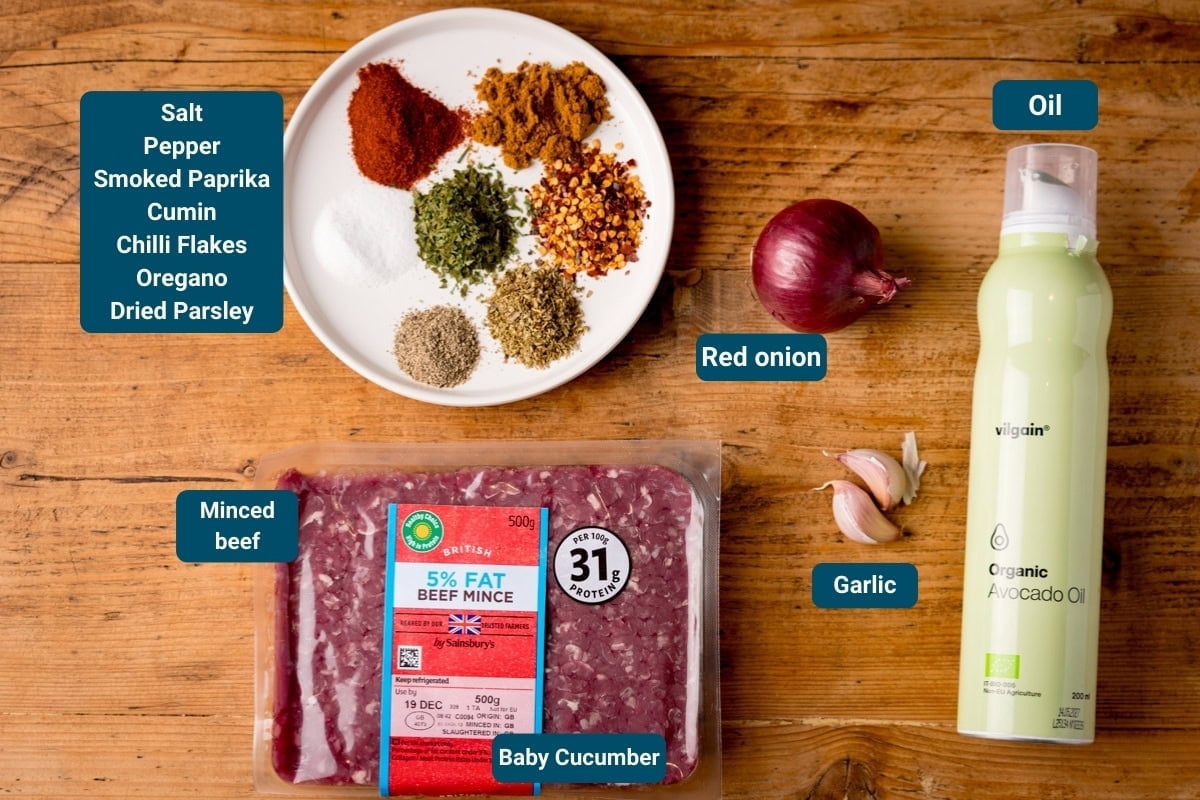 A wooden table shows labelled ingredients for Sheet Pan Kebabs: salt, pepper, smoked paprika, cumin, chilli flakes, oregano, dried parsley on a plate, red onion, oil, garlic, beef mince and baby cucumber.