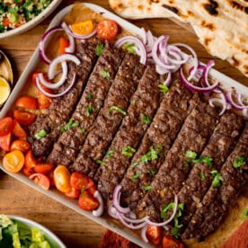 A tray of Sheet Pan Kebabs garnished with chopped parsley, red onion, and cherry tomato halves, served with flatbreads and salad on a wooden table.