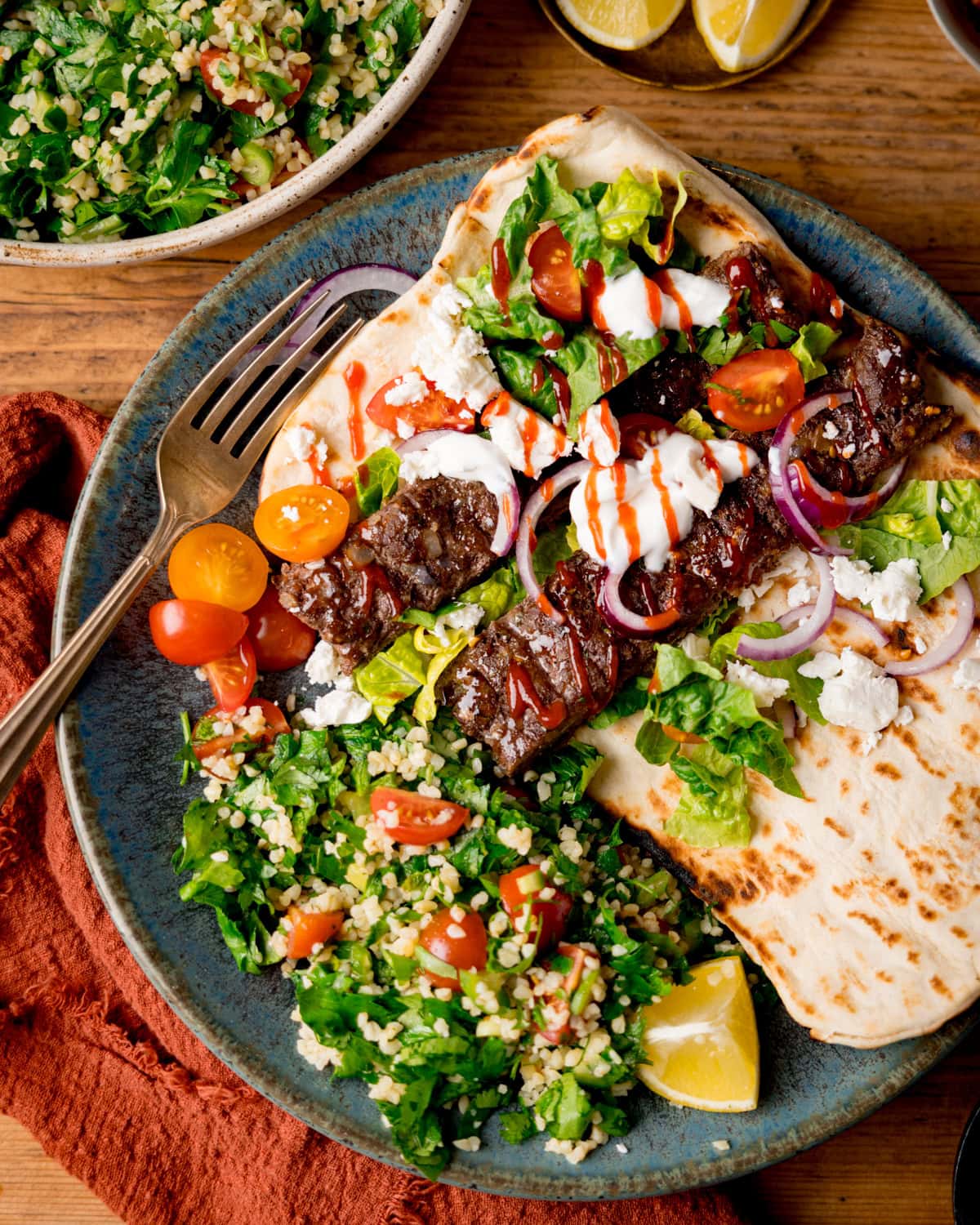 A blue-green plate with sheet pan kebabs on an open flatbread, topped with lettuce, tomatoes, onions, and sauces, served with tabbouleh salad and lemon wedges. The plate is on a wooden surface next to an orange napkin.