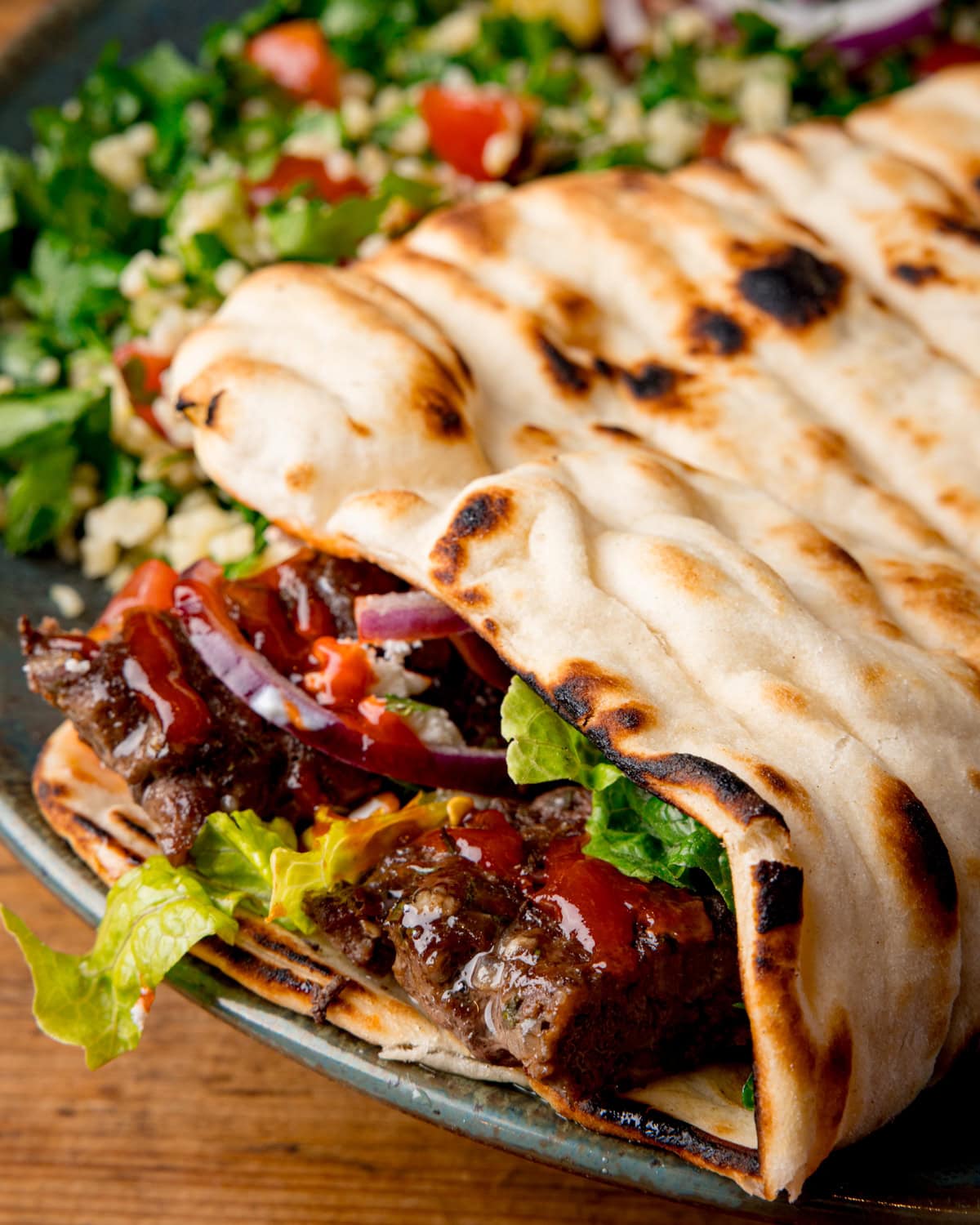 A close-up of grilled flatbread filled with sheet pan beef kebab, lettuce, red onion, and sauce. Served with tabbouleh salad on a blue-green plate.