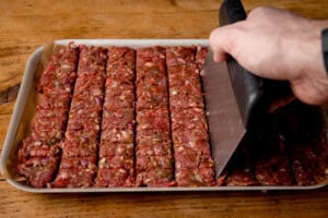 A hand uses a large bench scraper to score raw seasoned minced meat into rectangular strips on a baking paper-lined tray.