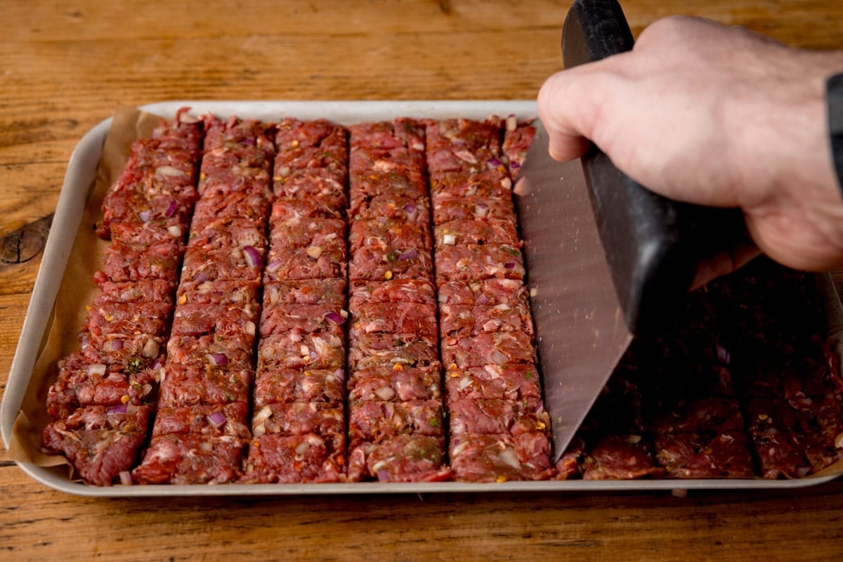 A hand uses a large bench scraper to score raw seasoned minced meat into rectangular strips on a baking paper-lined tray.