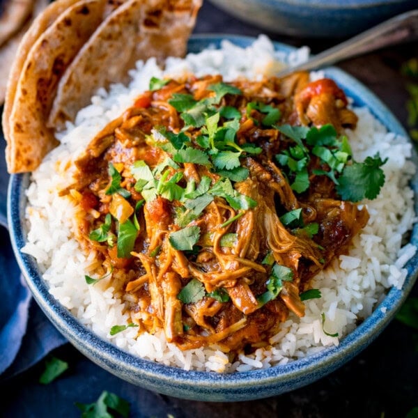 Square image of a blue bowl filled with white rice topped with slow cooked pulled lamb curry, garnished with coriander, served with flatbreads on the side.
