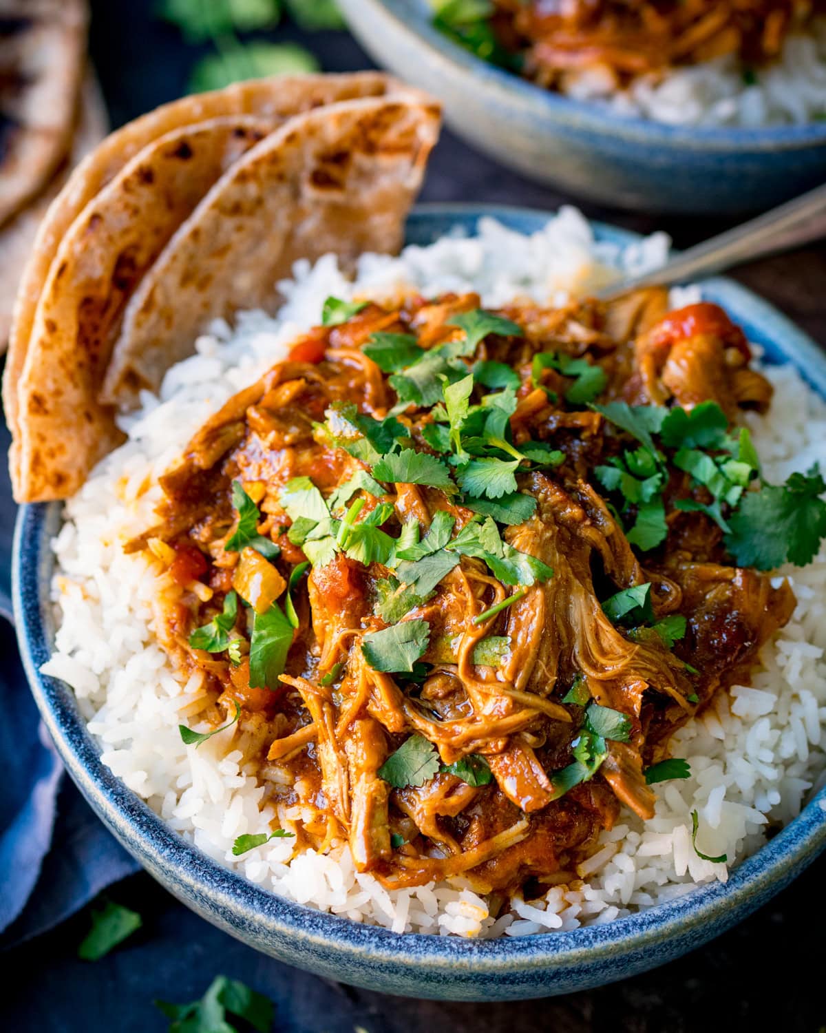 Tall image of a blue bowl filled with white rice topped with slow cooked pulled lamb curry, garnished with coriander, served with flatbreads on the side.