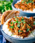Tall side-on image of a blue bowl filled with white rice topped with slow cooked pulled lamb curry, garnished with coriander, served with flatbreads on the side. There is a further bowl in the background.
