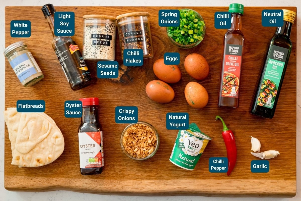A wooden board with labelled ingredients for spicy fried eggs - including: eggs, flatbreads, natural yoghurt, spring onions, white pepper, soy sauce, oyster sauce, sesame seeds, chilli flakes, crispy onions, garlic, chilli pepper and oils.
