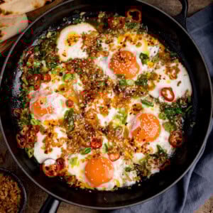 Square image of four sunny-side-up fried eggs in a skillet, topped with natural yogurt, fried onions, sliced red chiles, scallions, and sesame seeds. Some flatbreads are in the corner of the image.