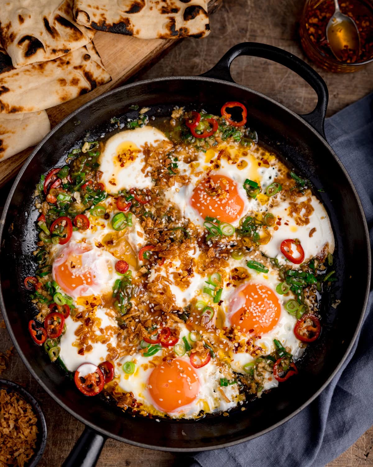 A frying pan of spicy fried eggs&mdash;four sunny-side-ups topped with chopped spring onions, red chilli slices, and crispy fried onions&mdash;served alongside pieces of flatbread.