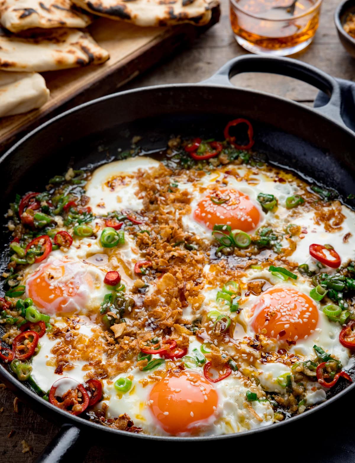Spicy Fried Eggs: Four sunny-side-up eggs cooked in a frying pan, topped with spring onions, red chilli slices and crispy fried onions, served with naan bread and a refreshing drink in the background.