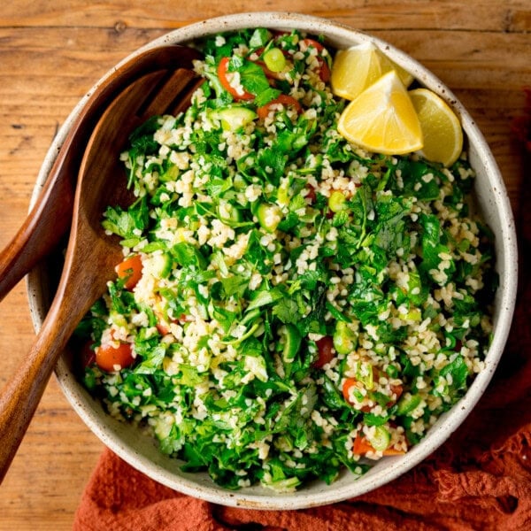 A bowl of tabbouleh salad with bulgur, parsley, tomatoes, and lemon wedges. Wooden salad are nestle in the bowl. The bowl sits next to an orange napkin, on a wooden table.