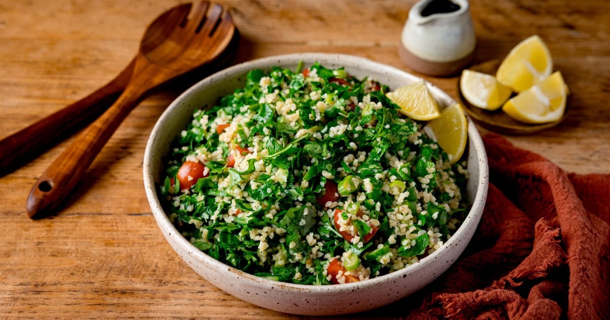 A bowl of chopped herbs, diced tomatoes and bulgur wheat with 2 small wedges of lemon.