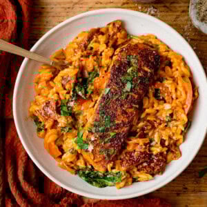 Square image of a white bowl with Tuscan-style orzo and pan fried salmon on top, garnished with parsley. The bowl is on a wooden table next to an orange napkin.
