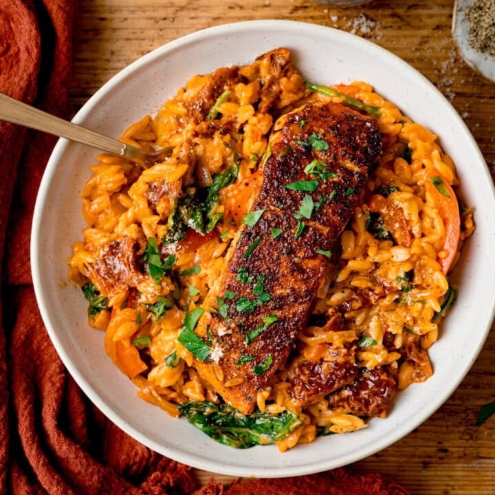 Square image of a white bowl with Tuscan-style orzo and pan fried salmon on top, garnished with parsley. The bowl is on a wooden table next to an orange napkin.