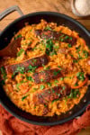 Overhead image of a dark pan filled with Tuscan-style orzo and four pan fried salmon fillets on top, garnished with parsley. There is a wooden spoon sticking out of the pan and the pan is next to an orange napkin. There is a small bowl of parmesan at the top of the image.