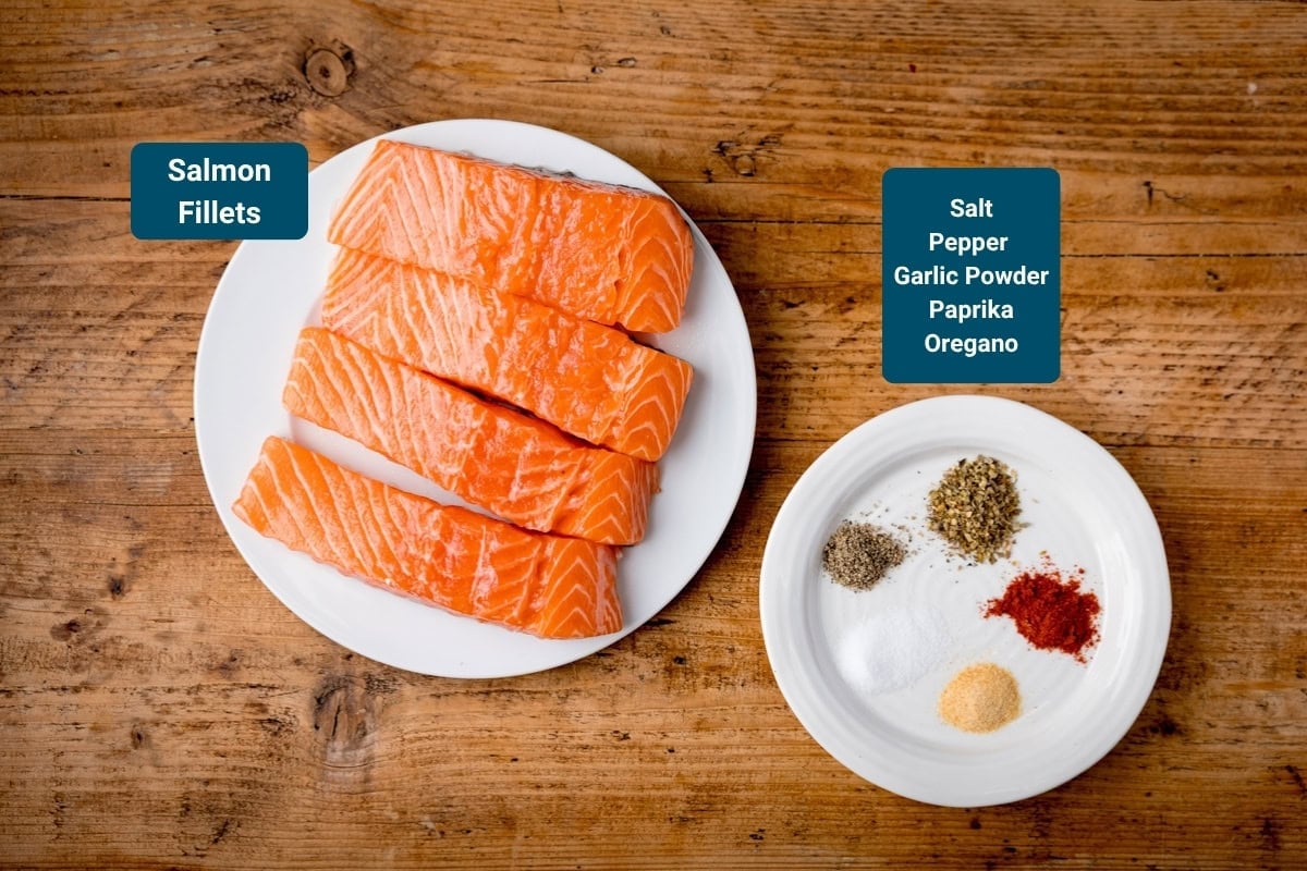 Four raw salmon fillets on a white plate sit next to a small white plate with seasonings on &mdash;salt, pepper, garlic powder, paprika, and oregano. The plates are on a wooden surface.