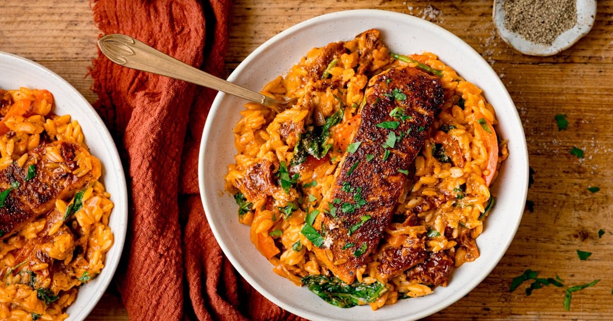 A white bowl with tomato flavoured orzo pasta with a crusted salmon fillet on top.