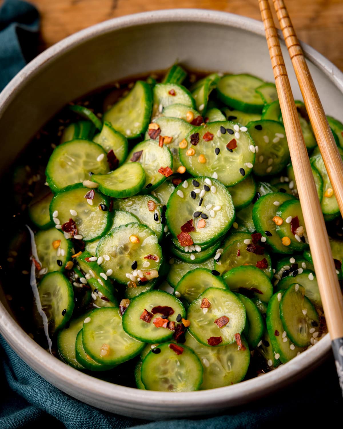 A bowl of Asian Cucumber Salad, with sliced cucumbers topped with sesame seeds, red chilli flakes, and a soy-sesame sauce, with a pair of chopsticks resting on the bowl.
