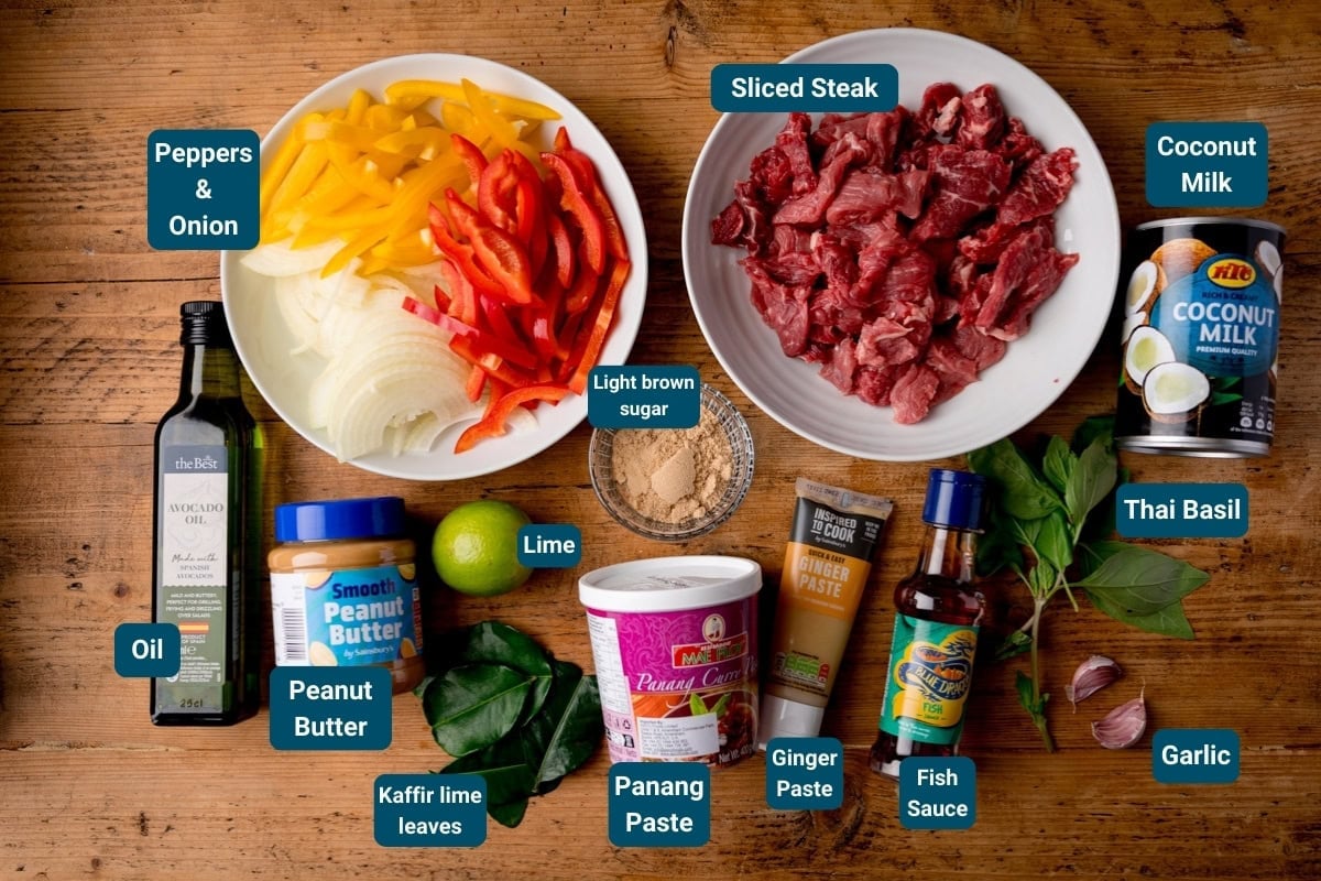 Top-down view of ingredients for a Beef Panang Curry, including sliced steak, peppers, onion, coconut milk, herbs, spices, sauces, and condiments arranged on a wooden surface.