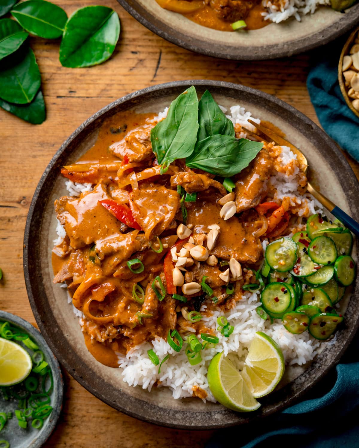 A plate of rice topped with creamy Beef Panang Curry, Asian cucumber salad, chopped peanuts, spring onions, lime wedges, and fresh Thai basil on a wooden table. Kaffir lime leaves, spring onions and part of a dark green napkin are also in shot.