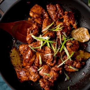 Chicken adobo in a dark glaze with bay leaves, topped with sliced spring onions, served in a black frying pan with a wooden spoon.