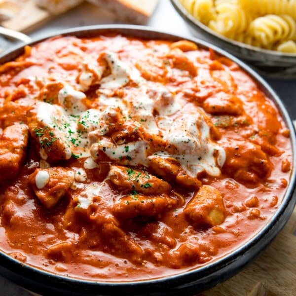 A close-up of Chicken Paprikash in a creamy tomato-based sauce, garnished with herbs, with a bowl of fusilli pasta in the background.