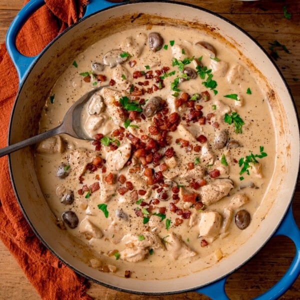 A blue cast iron pan filled with chicken in a cream-cider sauce, topped with crispy bacon and parsley. The pan sits on a wooden surface with an orange napkin.