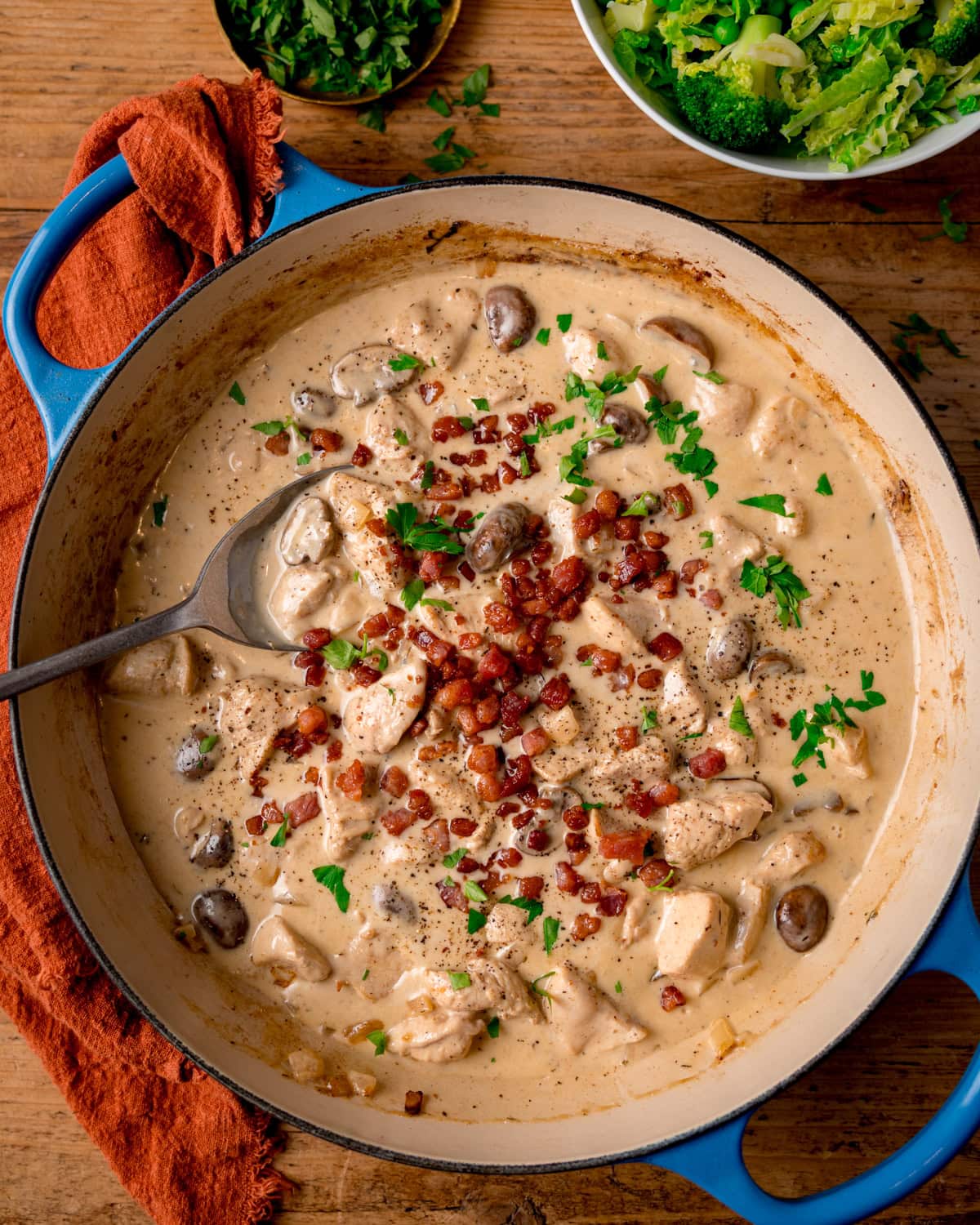 A blue cast iron pan filled with chicken in a cream-cider sauce, topped with crispy bacon and parsley. The pan sits on a wooden surface with an orange napkin. There is a bowl of green veg and a small bowl of parsley also in shot.