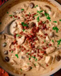 A close up overhead shot of chicken and mushrooms in a creamy cider sauce, topped with crispy bacon and parsley, in a large pan. A spoonful is being taken.