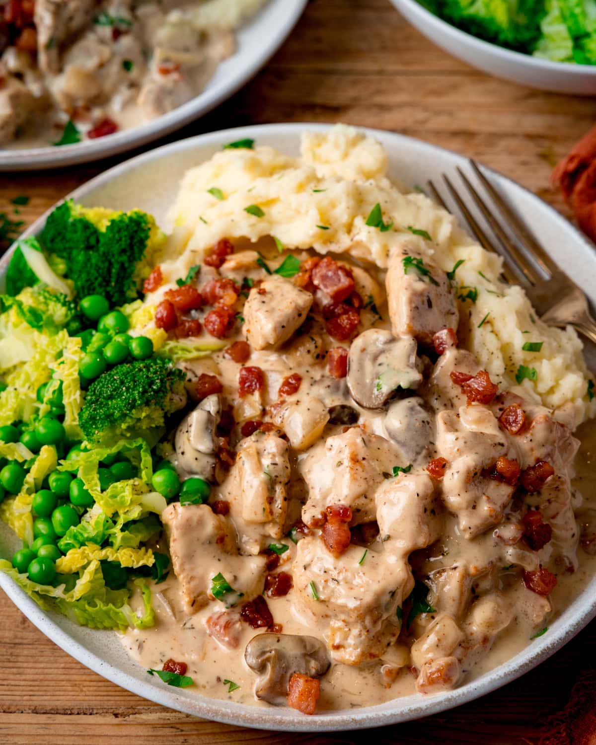 A plate of chicken in a creamy cider sauce with mushrooms and bacon, served with mashed potato, garden peas, broccoli, and cabbage.