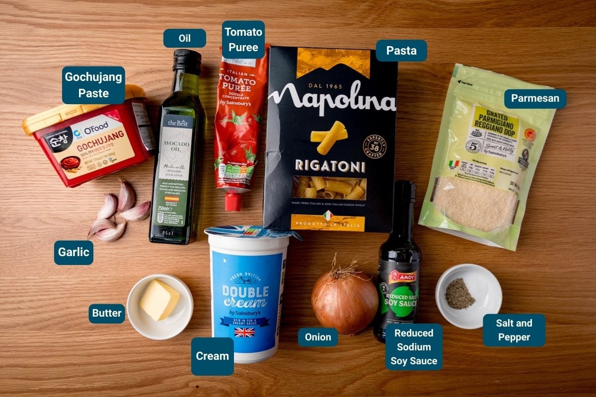 Ingredients for Gochujang Pasta on a wooden surface. All ingredients labelled, including rigatoni, cream, tomato pur&eacute;e, parmesan, gochujang paste, soy sauce, garlic, butter, onion, oil, salt and pepper.