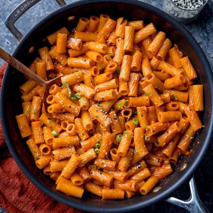 Overhead square image of a frying pan filled with creamy Gochujang pasta topped with grated Parmesan, black pepper, and chopped spring onions. There is a wooden spoon resting in the pan.