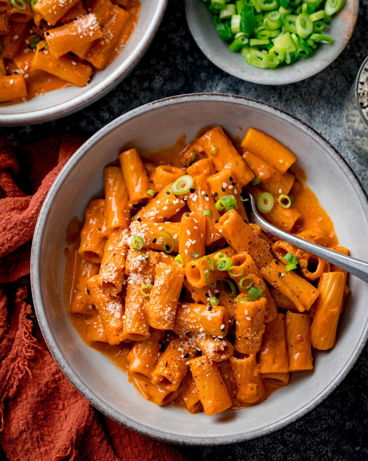 A bowl of spicy Gochujang pasta in a creamy gochujang sauce, topped with grated cheese, sliced spring onions, and black pepper, with a fork resting in the bowl. A further bowl of pasta and a dish of spring onions are also in shot.